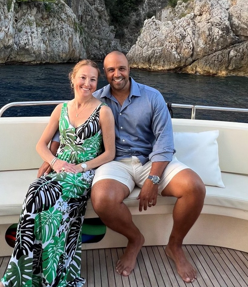 Ray and Rebecca Poole sitting close together on the back of a boat in Italy, smiling, with a rocky cove behind them.