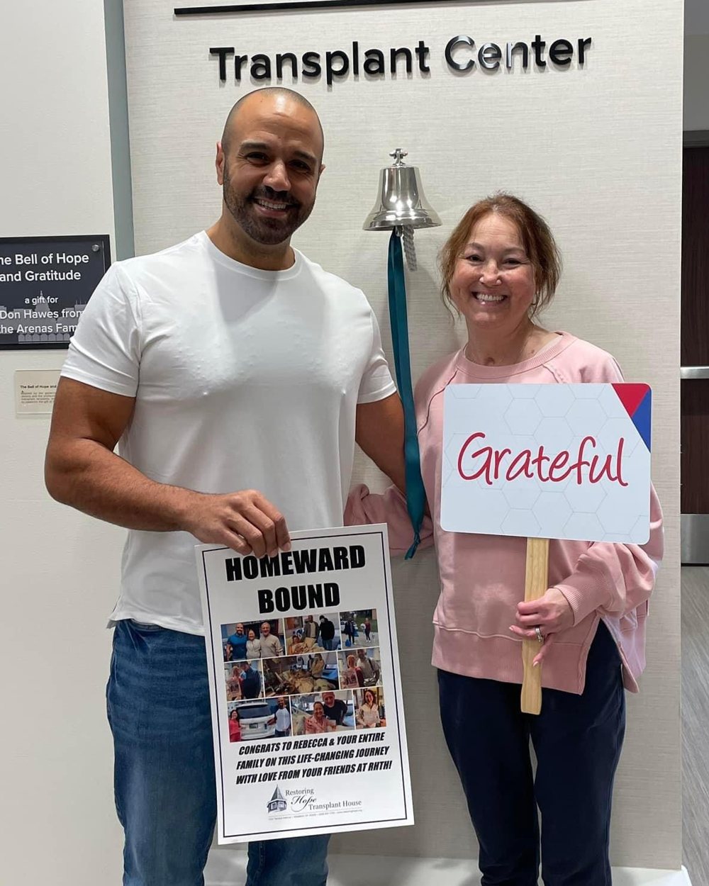 Ray and Rebecca smiling beside a hospital bell under a Transplant Center sign. Rebecca holds a sign that says Grateful.