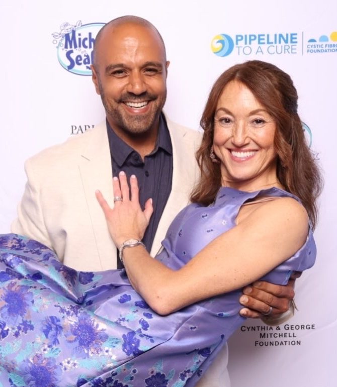 Ray Poole lifting Rebecca in his arms in front of a Pipeline to a Cure event backdrop, both laughing.