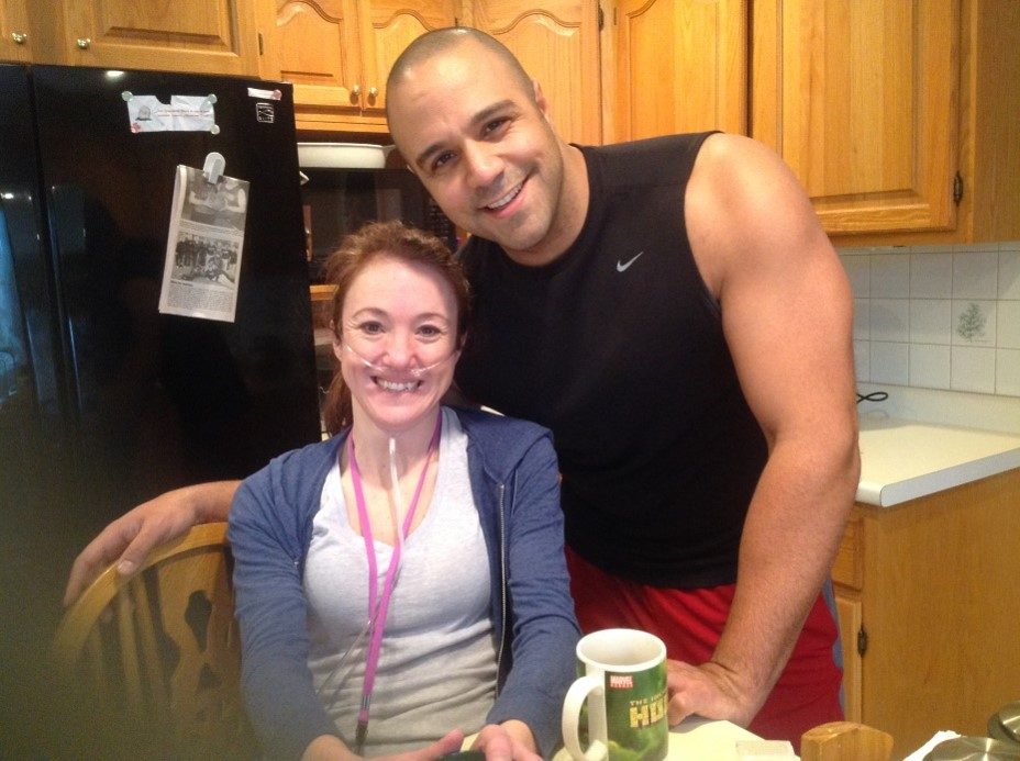 Rebecca smiling in a kitchen with a thin oxygen tube under her nose, and Ray leaning in behind her with a Hulk mug on the table.
