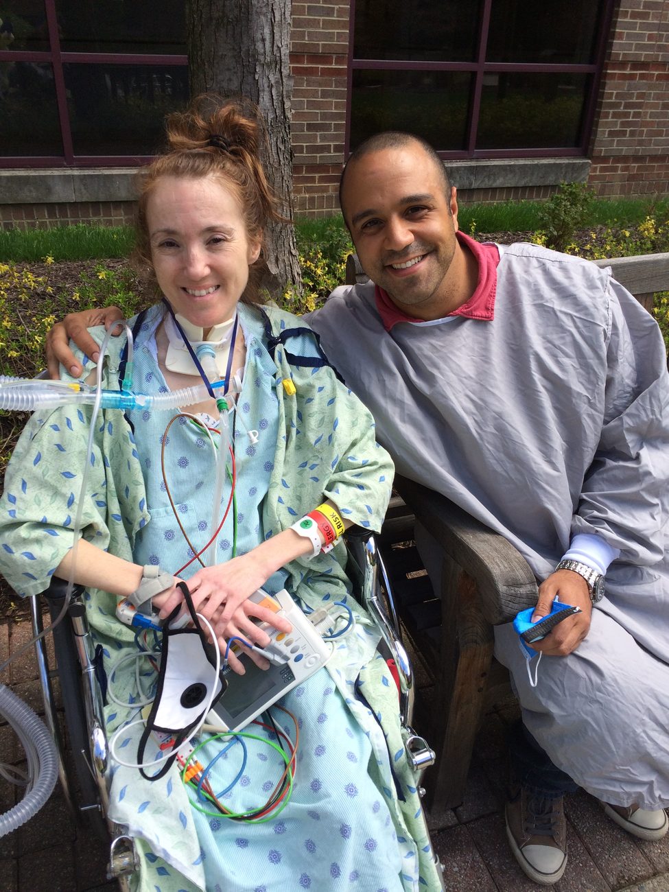 Rebecca in a wheelchair outdoors with a breathing tube and monitor, smiling beside Ray, who wears a hospital gown and sits on a bench.