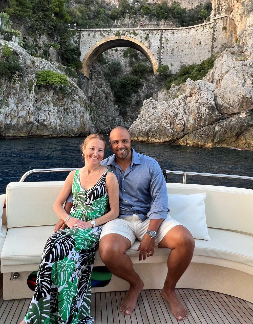 Ray and Rebecca sitting together on the back of a boat, with a tall stone bridge arching over a rocky cove behind them.