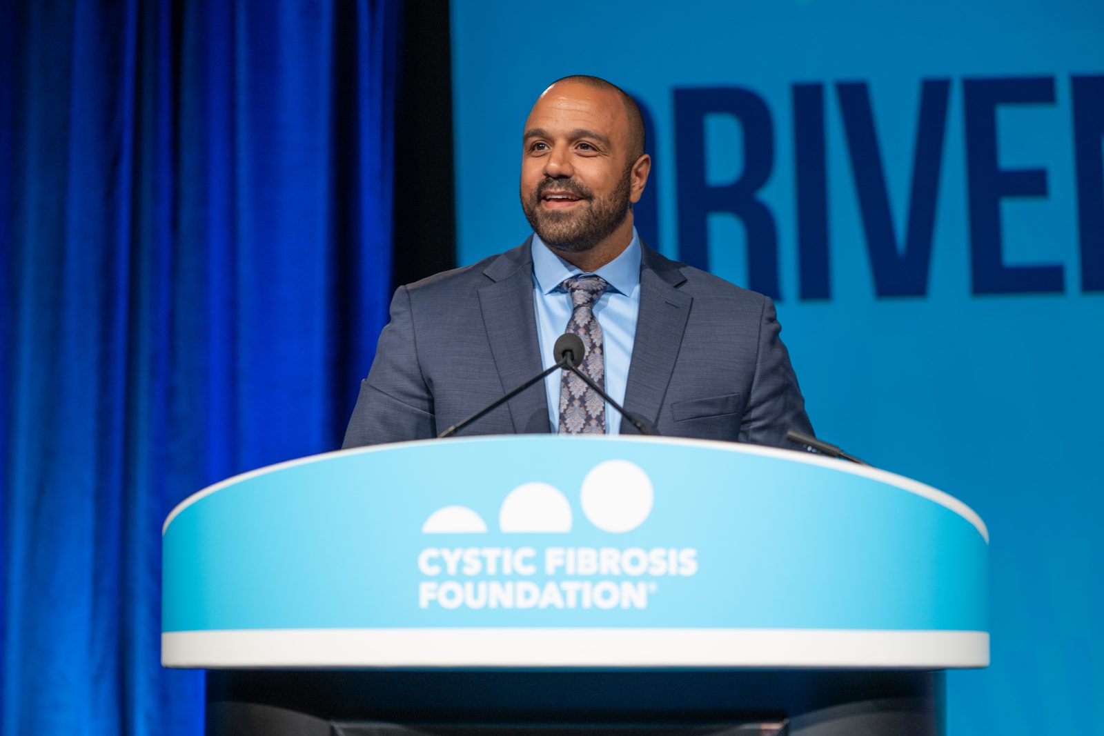 Ray speaking at a podium marked Cystic Fibrosis Foundation, smiling.